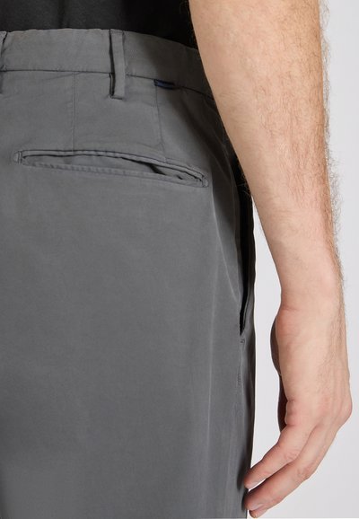 Primo piano del braccio di una persona accanto a pantaloni grigi con tasca posteriore orizzontale e passanti per cintura su uno sfondo semplice.