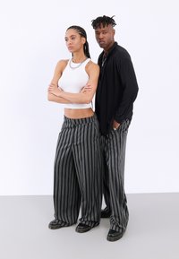 Două modele stau împreună, purtând pantaloni largi cu dungi alb-negru. Femeia are un top alb mulat, iar ambele poartă încălțăminte neagră.