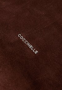 Tissu en daim marron avec un logo métallique argenté "COCCINELLE", positionné de manière proéminente à la surface avec une texture lisse.