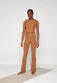 Barna crop top és testhezálló jóganadrág, mindkettő sima, rugalmas anyagból készült. A dizájn egyszerű, a felsőn egy logó található. Szőnyegen áll.