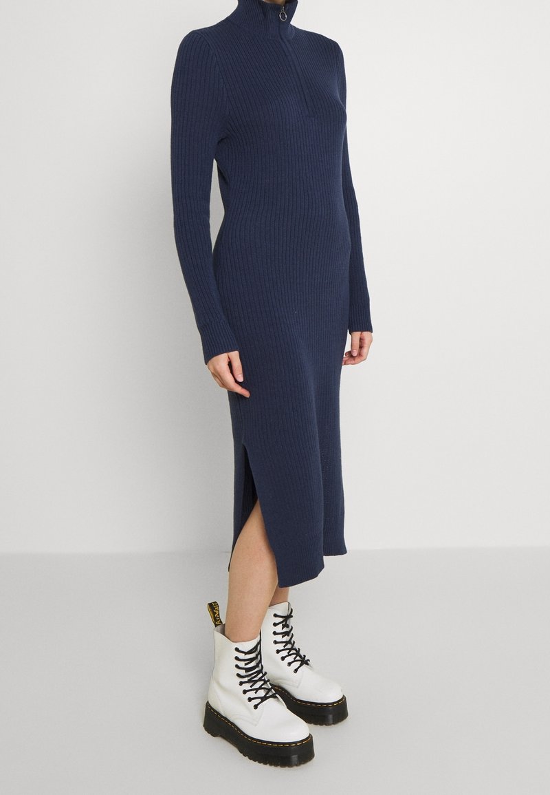 Femme portant une robe midi côtelée bleu marine avec une fente latérale et des bottines blanches à plateforme lacées, debout contre un mur uni.