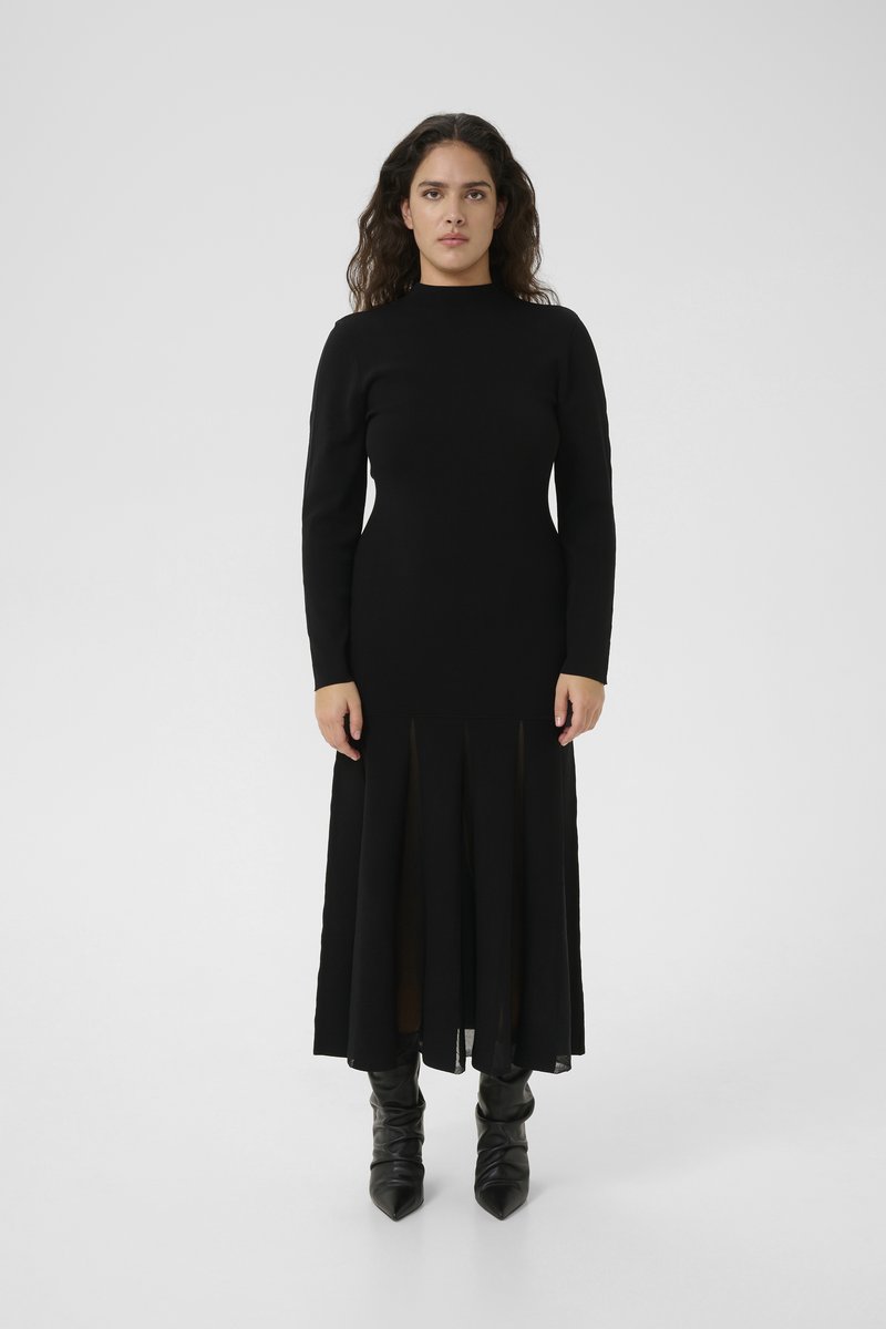 Schwarzes langärmliges Kleid mit hohem Ausschnitt, das eine gerade Silhouette und transparente Einsätze am Saum aufweist. Getragen mit kniehohen schwarzen Stiefeln.