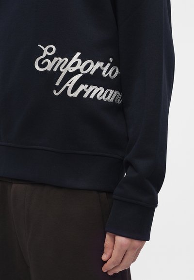 Chemise à manches longues foncée avec texte brodé blanc "Emporio Armani" sur le côté, associée à un pantalon foncé, montrant une main détendue sur le côté.
