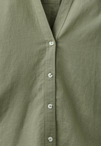 Chemise en lin vert olive clair avec col en V et quatre boutons blancs visibles à l'avant.