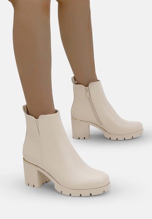 Bottines beiges avec une finition lisse, ressemblant à du cuir. Talons carrés et zip latéral avec des détails dorés. Semelle en caoutchouc texturé.