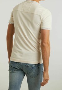 Beige t-paita lyhyillä hihoilla, jossa on yoke-muotoilu ja kontrastiväriset tikkaukset, yhdistettynä sinisiin farkkuihin, joissa on takataskut ja haalistuneita yksityiskohtia.