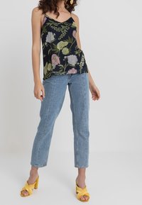 Kwiatowy top na ramiączkach na modelce, ciemnoniebieskie tło z różowymi i białymi kwiatami. Połączony z jasnoniebieskimi dżinsami i żółtymi sandałami na obcasie.