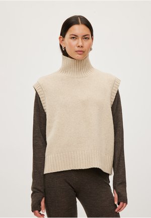 Femme portant un pull sans manches à col roulé beige sur un haut à manches longues marron foncé et un pantalon assorti, de face.