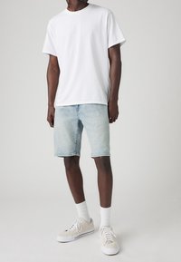 Muž oblečený v jednoduchém bílém tričku, světlých modrých džínových šortkách, bílých ponožkách a béžových plátěných teniskách stojí na světlejším pozadí.