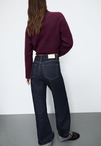 Temno modri široki traper hlače z visokim pasom, ki imajo svetlo šivanje in belo etiketo zadaj. Uparjene z maroonsko puloverjem.