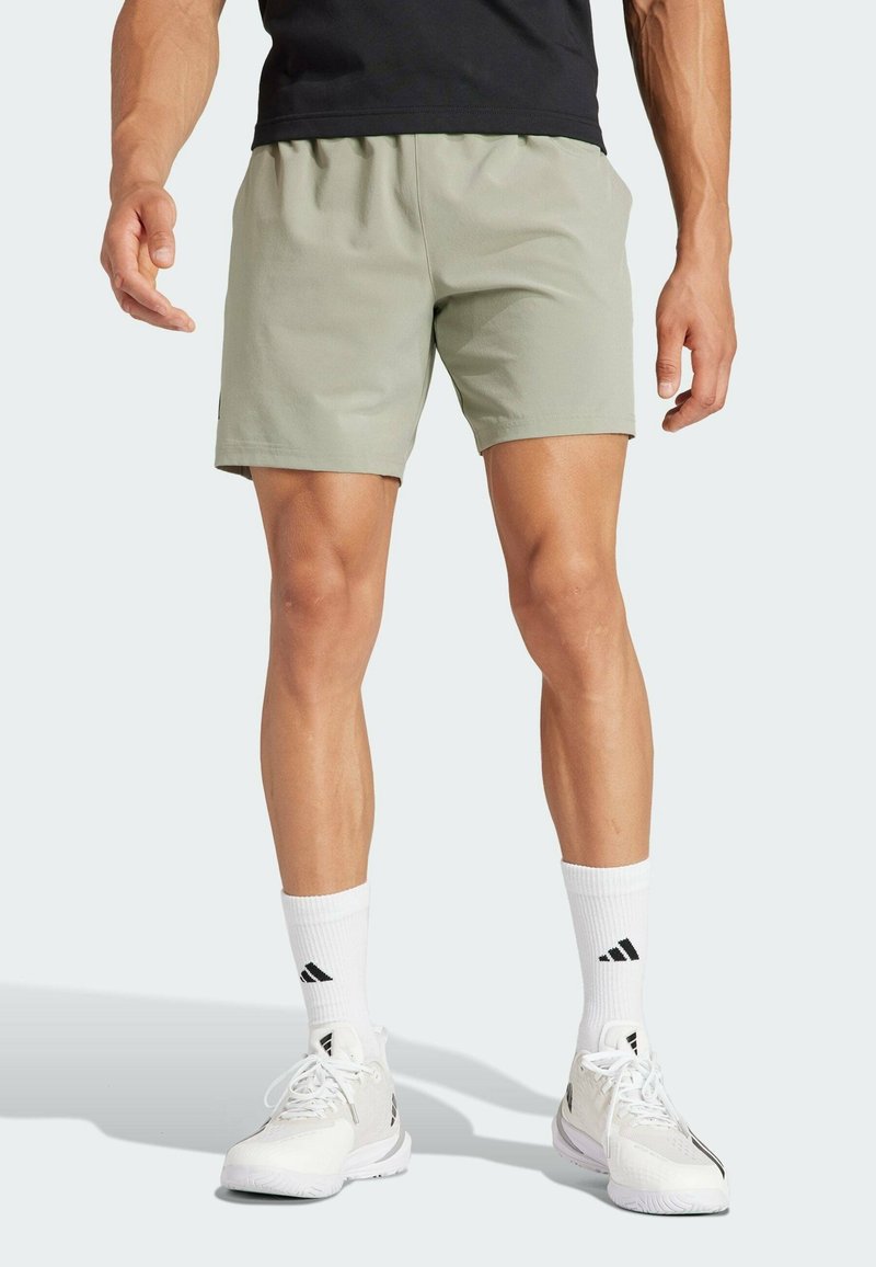 Homme portant un short de sport gris clair, des chaussettes blanches Adidas et des baskets blanches Adidas, debout devant un fond clair.