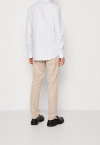 Persoon in beige broek, witte pinstripe shirt en zwarte leren schoenen, staande tegen een effen lichte achtergrond, met de rug naar de camera.