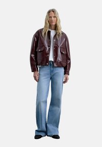 Veste en cuir bordeaux avec des fermetures à pression et des poches à rabat, associée à un jean évasé bleu clair et des chaussures noires, portée sur un fond uni.