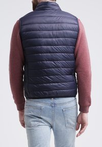 Námornická puffer vesta s horizontálním prošíváním, vysokým límcem a bez rukávů, nošena přes červený dlouhý rukáv, doplněná světle modrými džíny.