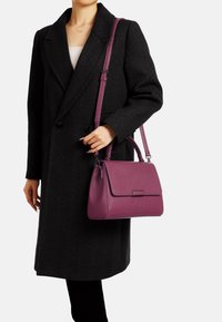 Borsa a tracolla in pelle viola con forma strutturata, chiusura a patta e tracolla removibile. È tenuta da una persona che indossa un cappotto nero.