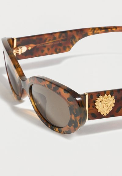 Οβάλ γυαλιά ηλίου με μοτίβο κέλυφος χελώνας, σκούρους φακούς και χρυσό έμβλημα σε σχήμα καρδιάς στην πλευρική βραχίωνα.