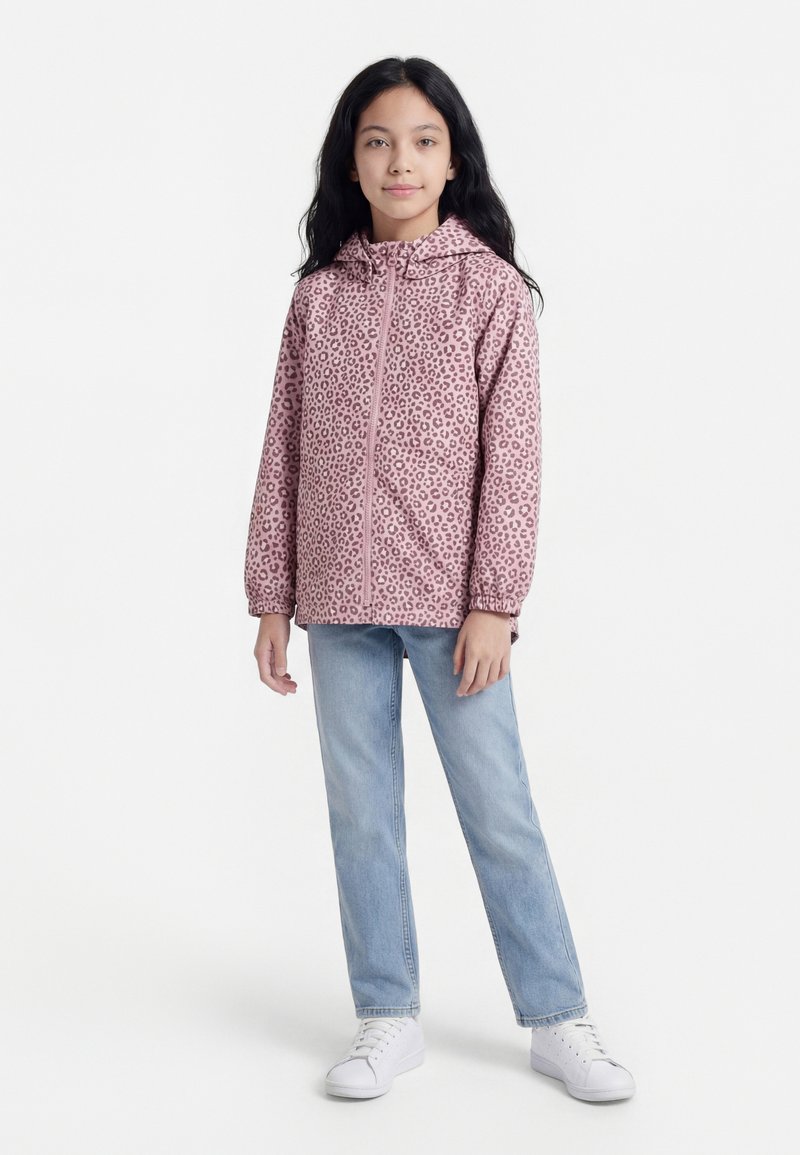 Fille debout portant une veste zippée rose à imprimé léopard, un jean bleu clair et des baskets blanches sur un fond blanc uni.