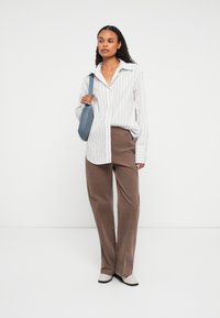 Vrouw in een witte gestreepte blouse, bruine corduroy broek, witte loafers en met een blauwe schoudertas, staande tegen een effen witte achtergrond.