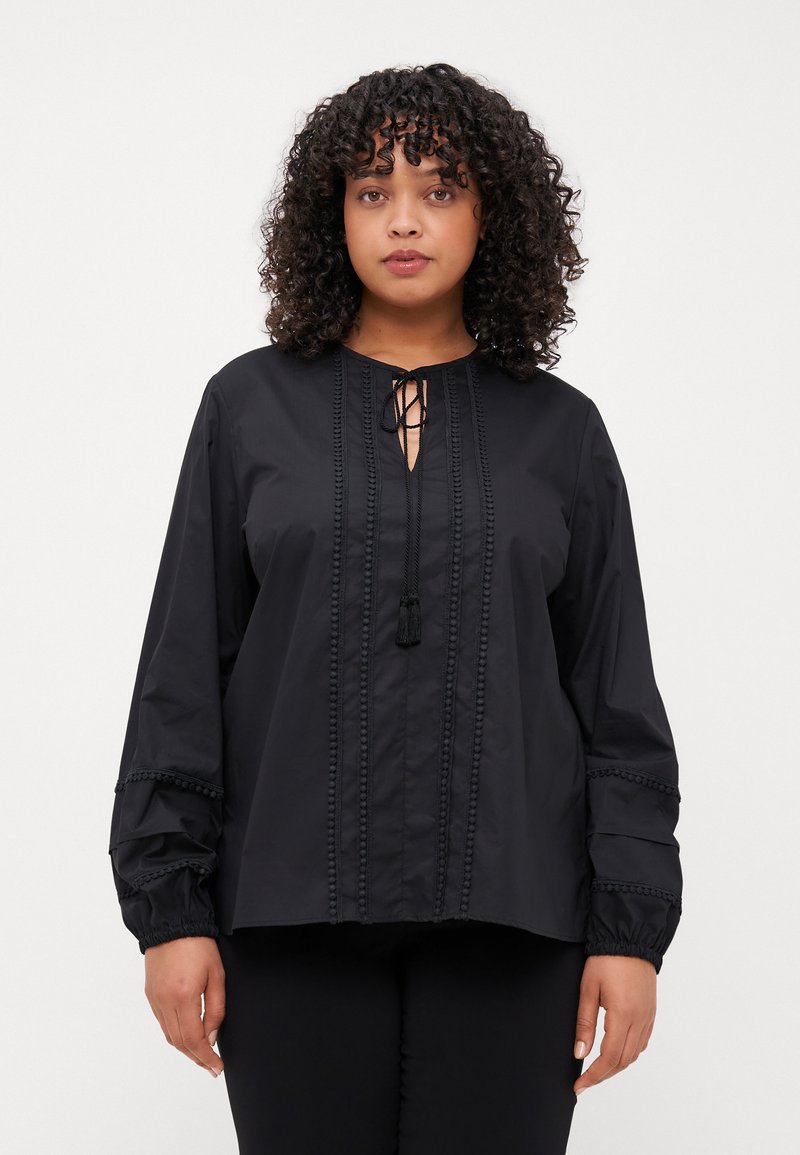 Svart skjorte med knapper og knyting i halsen, frontklaff, teksturert kant og elastiske ermer. Stoffet ser glatt ut med en lett glans.