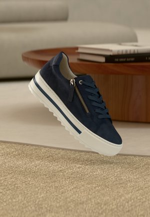 Marineblaue Wildleder-Sneaker mit weißer Sohle, ausgestattet mit Reißverschluss-Detail und Schnürung, auf einer strukturierten beigen Oberfläche präsentiert.