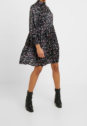 Mujer con vestido negro de flores, cuello alto, manga larga y botas negras hasta el tobillo sobre un fondo liso.
