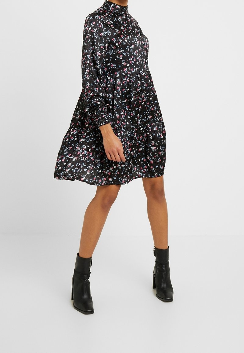Femme portant une robe noire à fleurs, col montant, manches longues et bottines noires sur un fond uni.