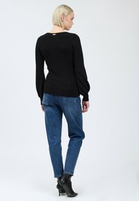 Donna con capelli biondi corti, che indossa una maglietta a maniche lunghe nera, jeans blu e stivaletti neri con tacco, rivolta di spalle su uno sfondo neutro.