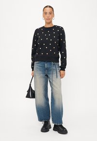 Sweatshirt noir avec des motifs de cœurs dorés, associé à un jean large bleu clair. Le modèle tient un petit sac à main noir et porte des bottes noires à lacets.