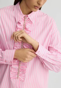 Blouse rose à rayures avec un devant volanté, des embellissements argentés le long du centre et des poignets avec des boutons roses.