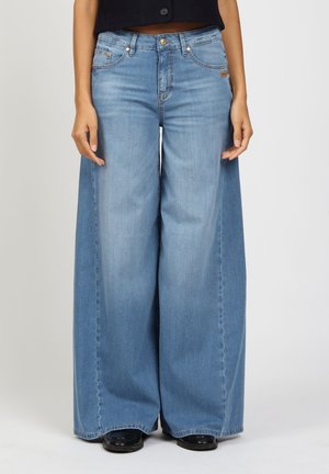 Woman wearing light blue wide-leg jeans and black shoes, standing with hands relaxed by sides against a plain background.