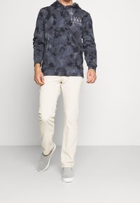 Felpa blu a camo con tasca a kangaroo frontale e logo, abbinata a pantaloni di velluto a coste beige e scarpe grigie slip-on, modello in cammino.