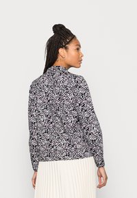 Blusa em preto e branco com padrões e detalhes em roxo, design ajustado, mangas longas e colarinho arredondado, usada sobre uma saia plissada em creme.