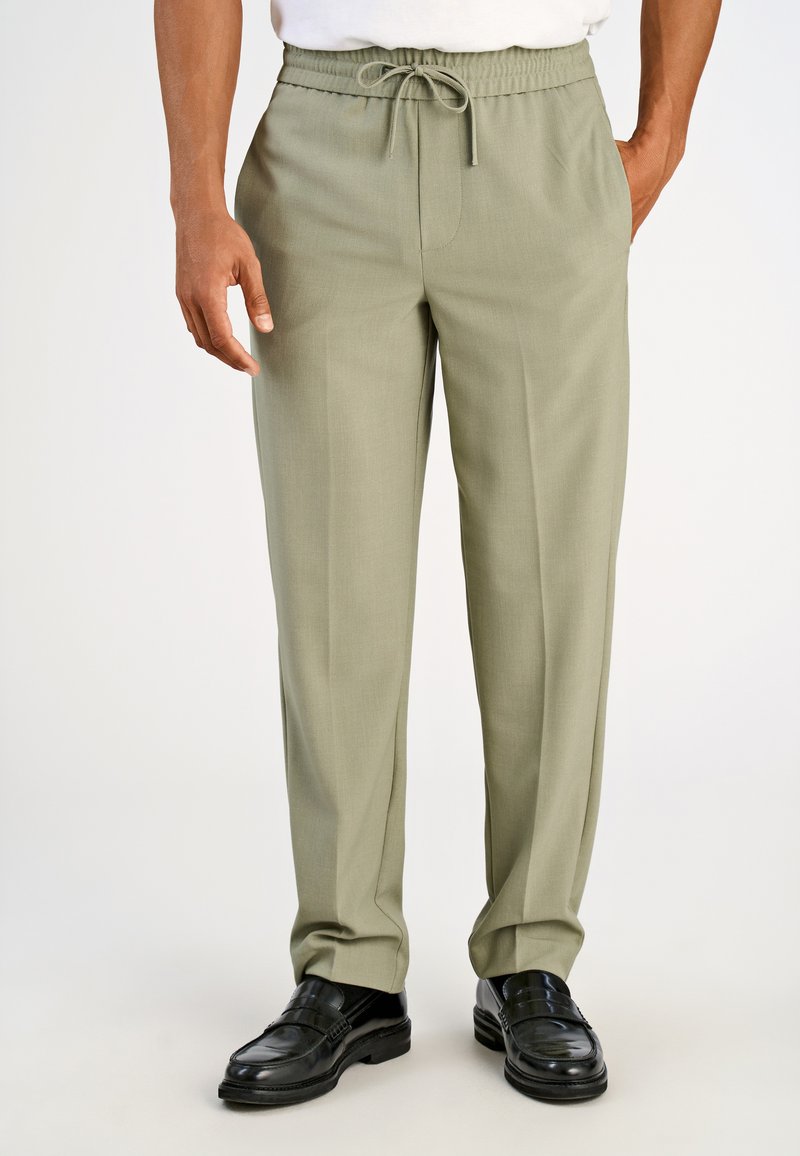 Uomo che indossa pantaloni con coulisse color oliva chiaro e mocassini di pelle neri, in piedi con le mani parzialmente nelle tasche su sfondo bianco.