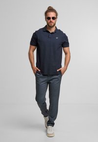 Polo shirt della marina con maniche corte, dettaglio logo sottile; pantaloni grigi texturizzati; accessoriumi con occhiali da sole rotondi e sneakers chiare.