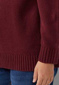 Bordeaux gebreide trui met een textuurlichaam en ribbelmanchetten. Zichtbare stiksels en een gladde, zachte afwerking van de stof. Hand rustend aan de zijkant.