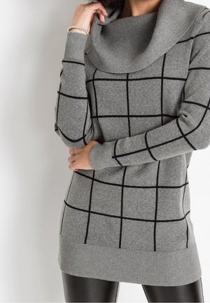 Woman wearing a gray knit sweater with black grid pattern and cowl neck, paired with black pants, standing with arms crossed and hand near face.