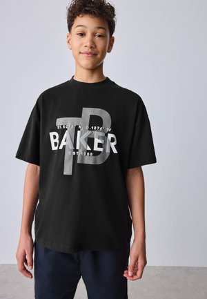 Teenager mit lockigem Haar trägt ein schwarzes oversized T-Shirt mit großem weißen und grauen „Baker“-Schriftzug und Koordinaten, stehend vor hellem schlichten Hintergrund.