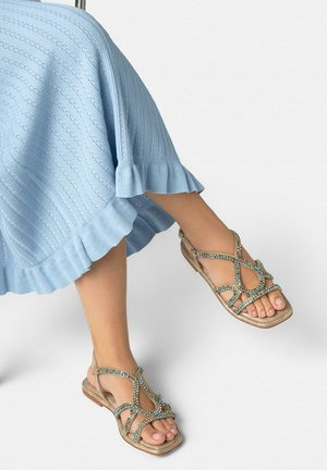 Pies con sandalias beige con piedras turquesas, combinadas con una falda de punto azul claro con dobladillo con volantes.
