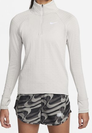 Kobieta ubrana w jasnoszary, rozpinany na zamek, długi rękaw z otworami na kciuki oraz czarno-szare, wzorzyste szorty Nike, stojąca na białym tle.