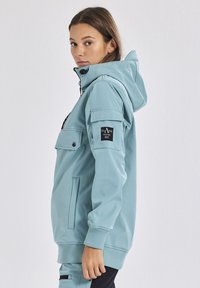 Giacca a vento azzurra chiara con cappuccio e tasca frontale, polsini elasticizzati e dettaglio con toppa sulle spalle. Texture liscia, vestibilità rilassata e materiale resistente.
