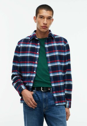 Junger Mann trägt ein blau-rotes-weiß kariertes Hemd über einem grünen T-Shirt mit blauen Jeans und schwarzem Gürtel, steht vor einem weißen Hintergrund.