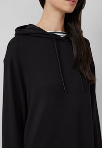 Schwarzer Kapuzenpullover mit lockerem Schnitt, Kordelzug und weicher Textur. Sichtbares gestreiftes Muster unter dem Kragen.
