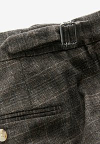 Grau karierte Hose aus einem Wollmischgewebe mit strukturierter Oberfläche, ausgestattet mit Gürtelschlaufen und einem metallischen Schnallenakzent. Sichtbares Detail der Vordertasche.