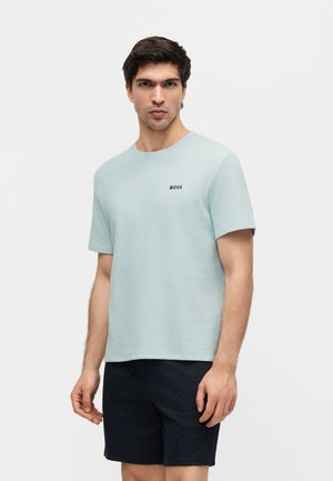 Férfi, aki világoskék rövid ujjú inget visel "BOSS" logóval és fekete rövidnadrágot, egy egyszínű fehér háttér előtt áll.