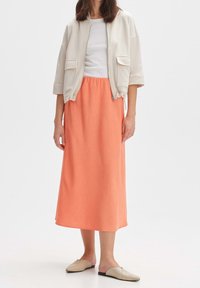 Giacca bomber grigio chiaro con maniche corte e due tasche anteriori, abbinata a una lunga gonna corallo strutturata e a delle scarpe slip-on beige.