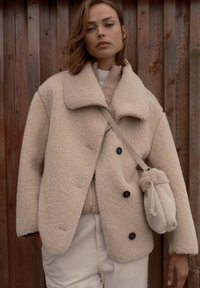 Beige, flauschiger Lammfellmantel mit breitem Kragen, doppelreißigen Knöpfen, kombiniert mit einer beigen Handtasche aus weichem Material.