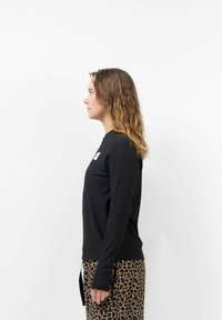 Schwarzes Langarmshirt mit geripptem Design und seitlichem Schlitzsaum, kombiniert mit Hose im Leopardenmuster. Modell im Profil gezeigt.