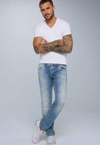 Vaqueros azul claro con un ajuste entallado, aspecto desgastado y costuras visibles, combinados con una camiseta blanca de corte en V ceñida y zapatillas blancas.