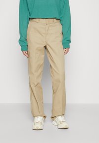 Pantaloni in cotone beige con taglio dritto e passanti per cintura, abbinati a una camicia verde petrolio a maniche lunghe e sneakers bianche.