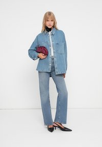 Mulher a usar uma jaqueta de denim oversized e jeans, segurando uma pequena mala roxa, sapatos de salto agudo pretos e um acessório preto para o pescoço.
